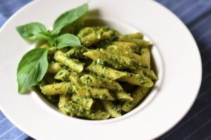 Penne pasta tossed in vibrant green basil pesto, garnished with fresh basil leaves, served in a white bowl on a blue tablecloth.