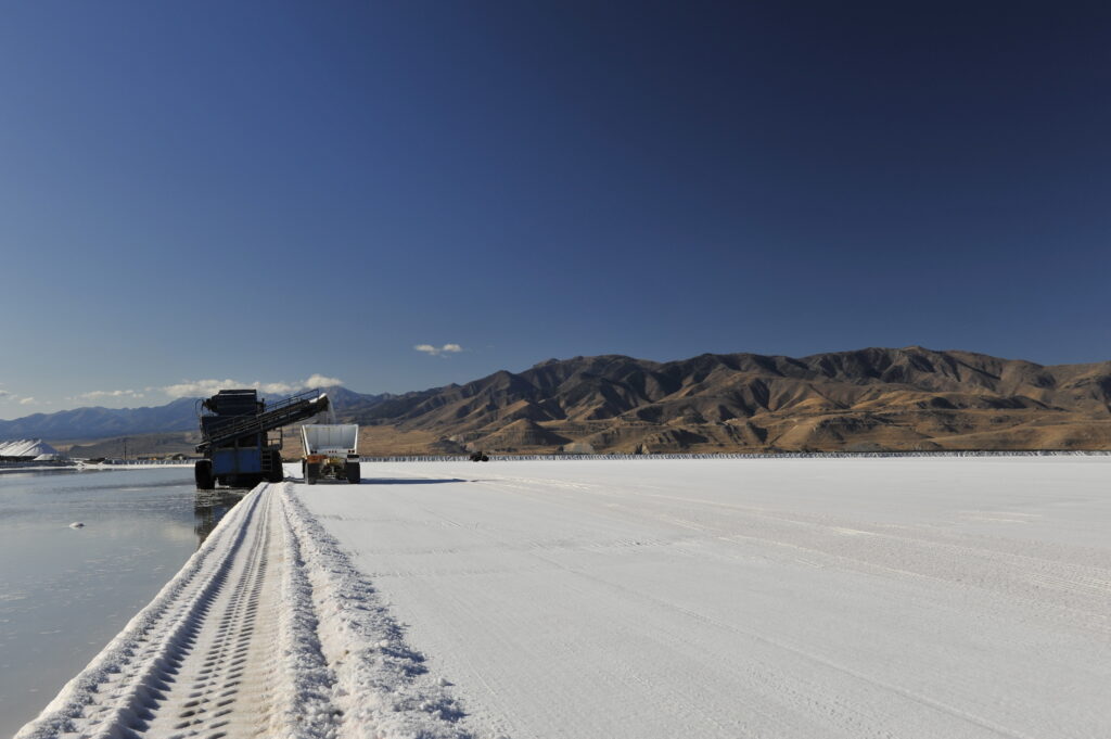 Heavy machinery harvesting salt from a large salt flat, with mountains in the background and a clear blue sky. The scene showcases the industrial process of salt extraction in a natural landscape.