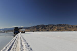 Heavy machinery harvesting salt from a large salt flat, with mountains in the background and a clear blue sky. The scene showcases the industrial process of salt extraction in a natural landscape.