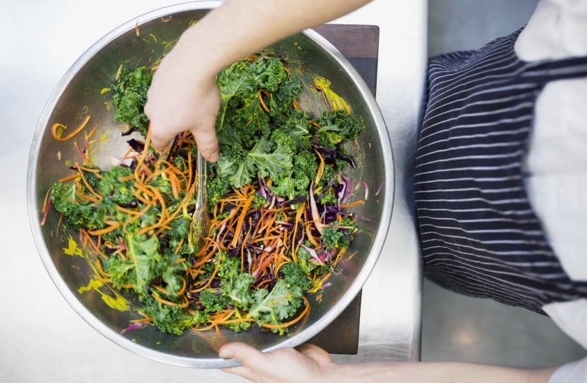 A chef's hands mix a vibrant salad of kale, purple cabbage, and shredded carrots in a stainless steel bowl, showcasing fresh ingredients in a professional kitchen setting.