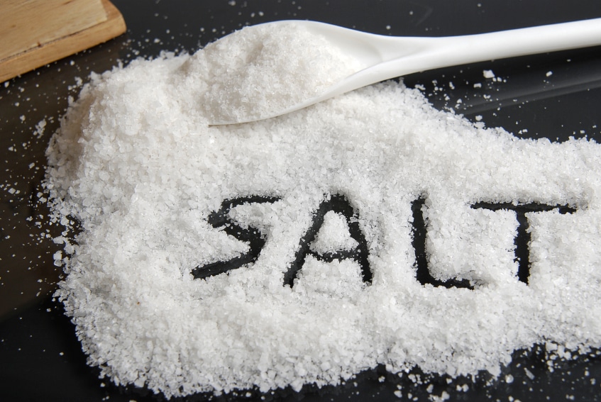 Close-up of coarse sea salt with the word "SALT" written in the salt, accompanied by a white spoon, highlighting the texture and purity of the salt on a dark surface. Perfect for culinary and cooking-related content.