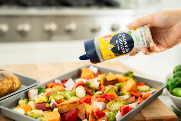 A hand pouring Morton Nature's Seasons seasoning blend over a tray of colorful roasted vegetables, including Brussels sprouts, bell peppers, and sweet potatoes, with a blurred kitchen background.