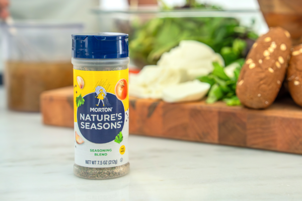 Morton Nature's Seasons seasoning blend bottle on a kitchen countertop, surrounded by fresh ingredients like salad greens and mushrooms, showcasing its versatile use in cooking.