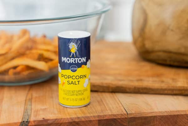Morton Popcorn Salt container on a wooden surface with a glass bowl of snacks in the background, highlighting its use for flavoring popcorn and other snacks.