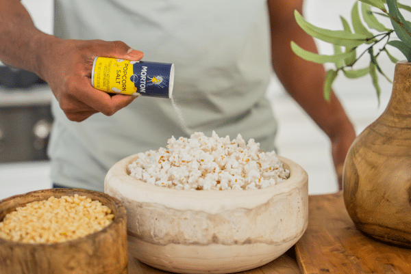 Person seasoning a bowl of freshly popped popcorn with Morton popcorn salt, with a wooden bowl of popcorn kernels in the background. Perfect for movie nights or snacking.