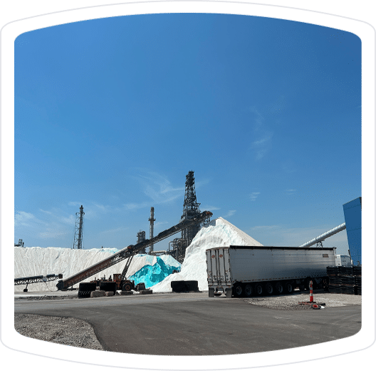 Industrial site with large salt piles and conveyor systems under a clear blue sky, featuring heavy machinery and a trailer truck in the foreground.