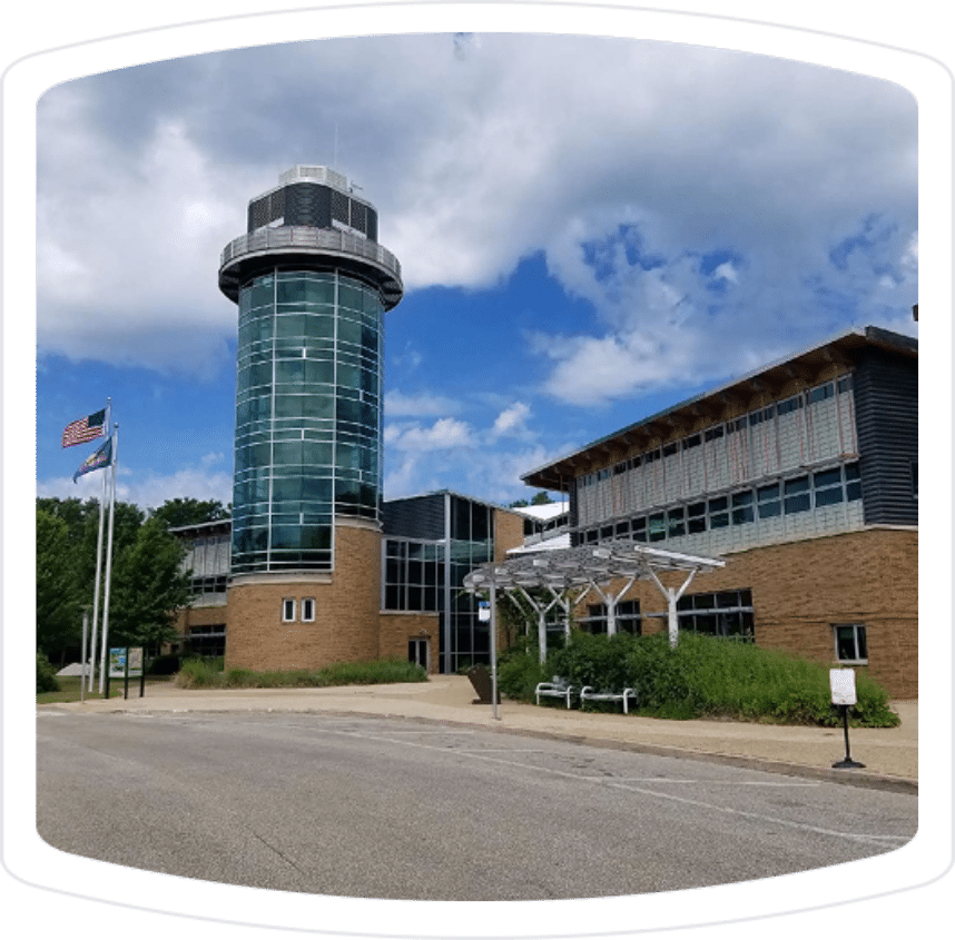 Modern building with a glass tower and eco-friendly design, featuring American and state flags, surrounded by greenery and a paved pathway. Ideal for showcasing sustainable architecture and community spaces.