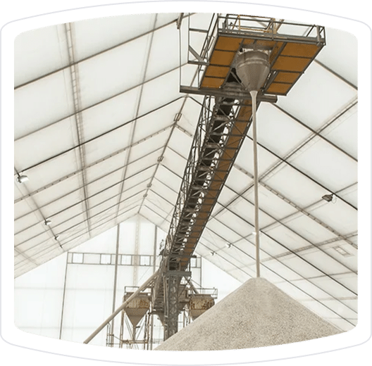 Conveyor system for transporting materials inside a large industrial facility, featuring a hopper and a pile of sand or aggregate below. The structure is covered by a translucent roof, allowing natural light to illuminate the workspace.