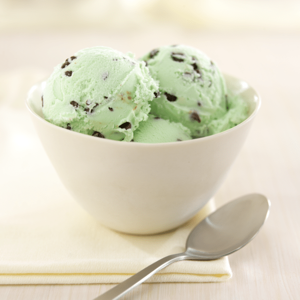 Bowl of mint chocolate chip ice cream with three scoops, placed on a yellow napkin beside a silver spoon, set against a soft, neutral background.