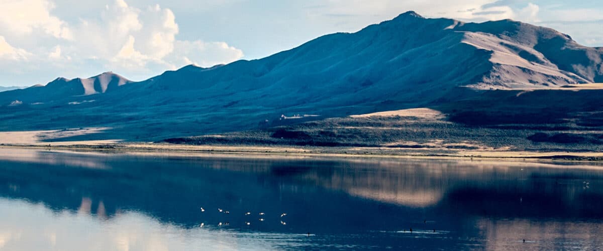 Scenic view of mountains reflecting in a calm lake under a blue sky, featuring a serene landscape with gentle slopes and scattered clouds.