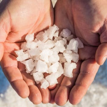 Close-up of two hands holding coarse sea salt crystals, showcasing their texture and size against a blurred background of salt. Ideal for topics on cooking, natural ingredients, or health benefits of salt.