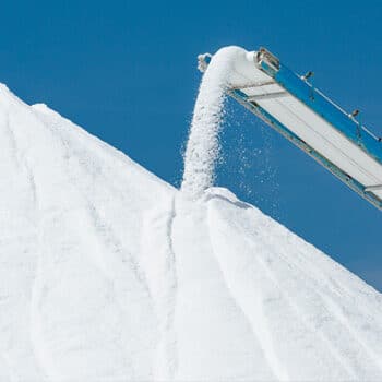 Conveyor belt pouring white salt onto a large pile against a clear blue sky, highlighting industrial salt production and storage.