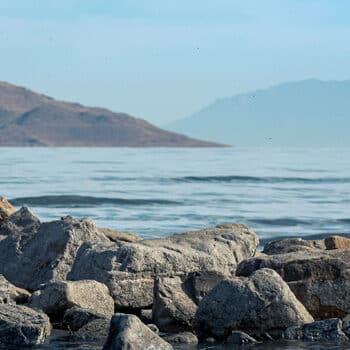 Rocky shoreline with calm water and distant mountains under a clear sky. Ideal for nature enthusiasts and landscape photographers.