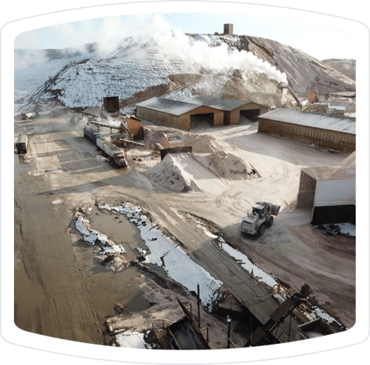 Aerial view of an industrial site featuring large storage facilities, trucks, and a mountain of material with steam rising, indicating an active processing operation. The landscape shows dirt roads and piles of raw materials, typical of a mining or construction area.