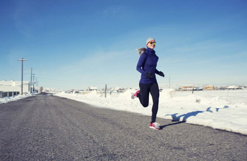Woman running on a snow-covered road in winter attire, wearing sunglasses and athletic gear, with a clear blue sky in the background.
