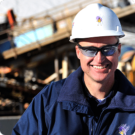 Smiling construction worker wearing a hard hat and safety glasses, standing in front of a construction site with machinery in the background.
