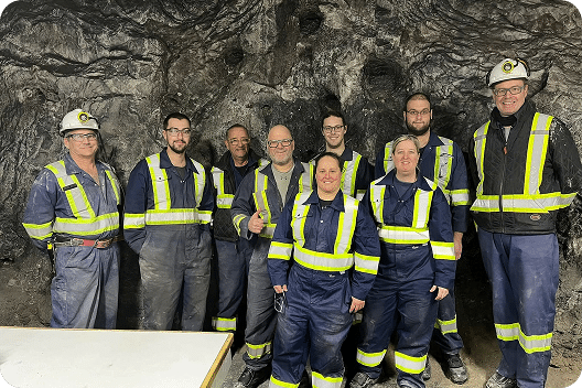 Group of nine mining professionals in safety gear, including hard hats and reflective vests, posing in a mine environment with rocky walls in the background. The team is engaged in mining operations, showcasing teamwork and safety in the industry.