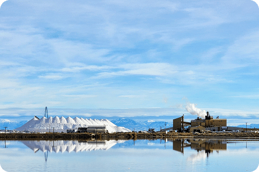 Salt production facility with large salt piles and industrial buildings reflected in calm water under a clear blue sky.