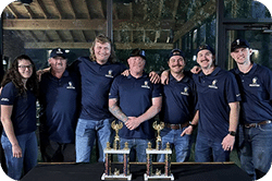 Group of seven people in navy shirts celebrating with trophies at an awards ceremony.