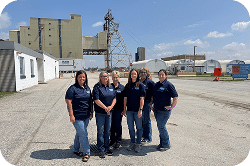 Group of six women in navy blue shirts standing outdoors at an industrial site, with grain silos and machinery in the background, showcasing teamwork and professionalism in a rural setting.
