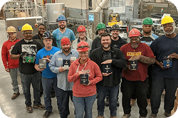 Group of factory workers wearing safety helmets and holding awards, posing together in a manufacturing facility. The image showcases teamwork and recognition in an industrial setting.