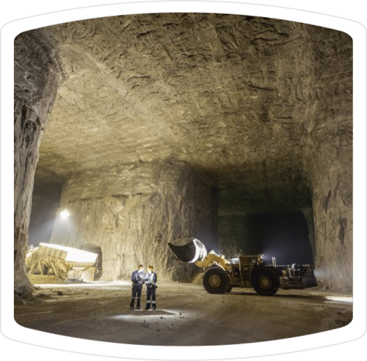 Two workers in safety gear stand in a large underground mining cavern, illuminated by artificial lights, with heavy machinery, including a loader, visible in the background. The cavern walls are made of rock, showcasing the scale of the mining operation.