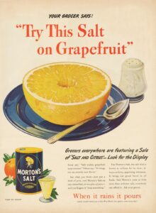 Vintage advertisement featuring a halved grapefruit on a blue plate, accompanied by a silver spoon and a salt shaker, promoting the use of Morton's Salt on citrus fruits. The ad highlights the flavor enhancement that salt brings to grapefruit and other citrus, encouraging customers to try this combination.
