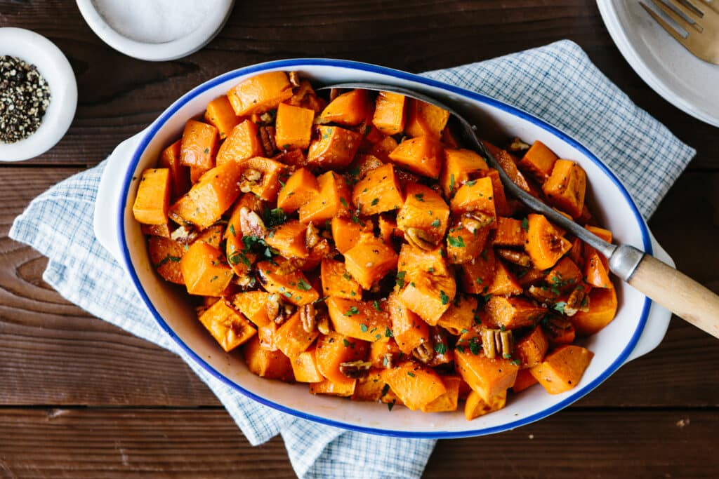 Bowl of roasted butternut squash cubes mixed with chopped pecans and garnished with fresh herbs, served on a blue and white checkered cloth.