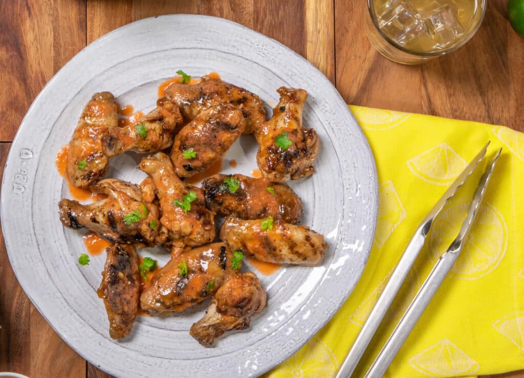 Grilled chicken wings coated in a savory sauce, garnished with fresh cilantro, served on a rustic plate with a yellow napkin and tongs in the background. Perfect for a barbecue or party appetizer.