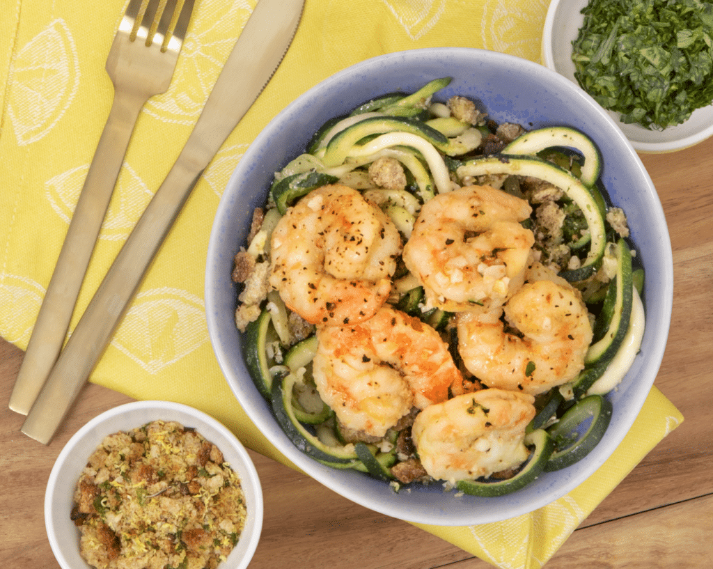 Shrimp and zucchini noodle dish served in a blue bowl, garnished with breadcrumbs and fresh herbs, accompanied by a small bowl of seasoned breadcrumbs, set on a bright yellow lemon-patterned napkin with gold utensils.
