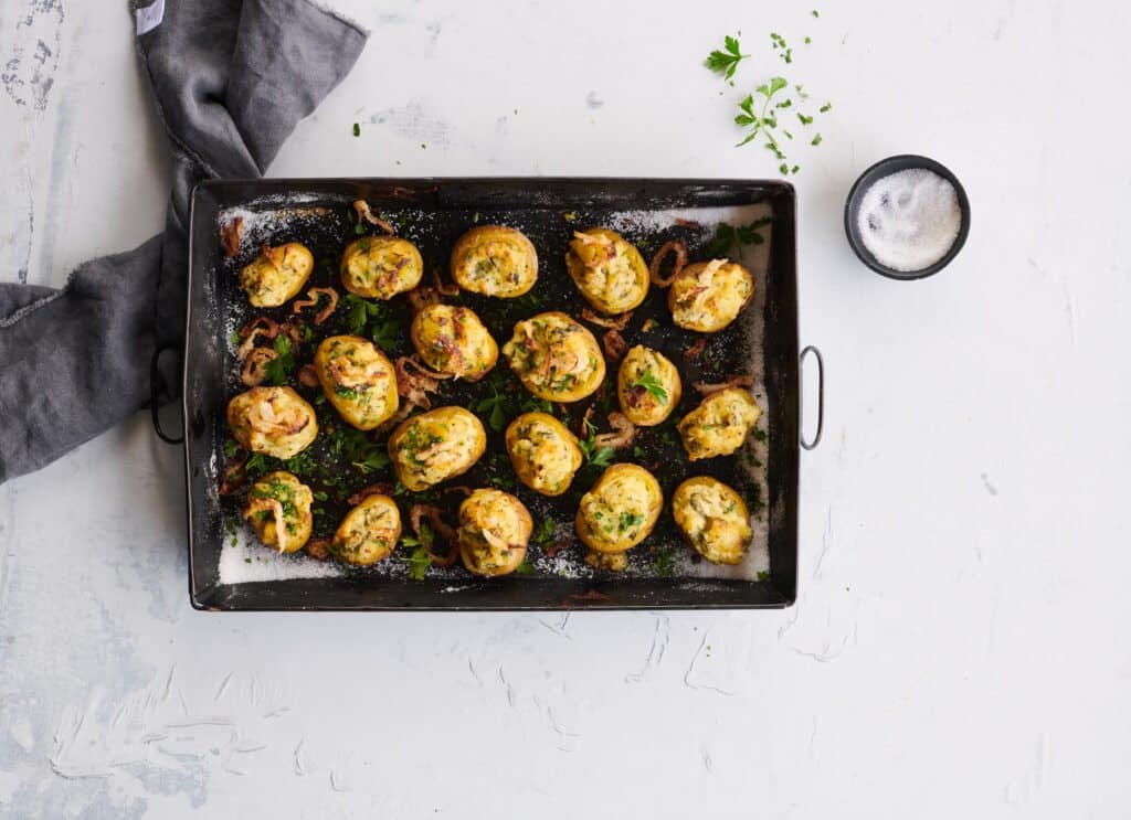 Baked stuffed potatoes topped with crispy onions and fresh parsley on a black baking tray, accompanied by a small bowl of salt, against a light textured background.