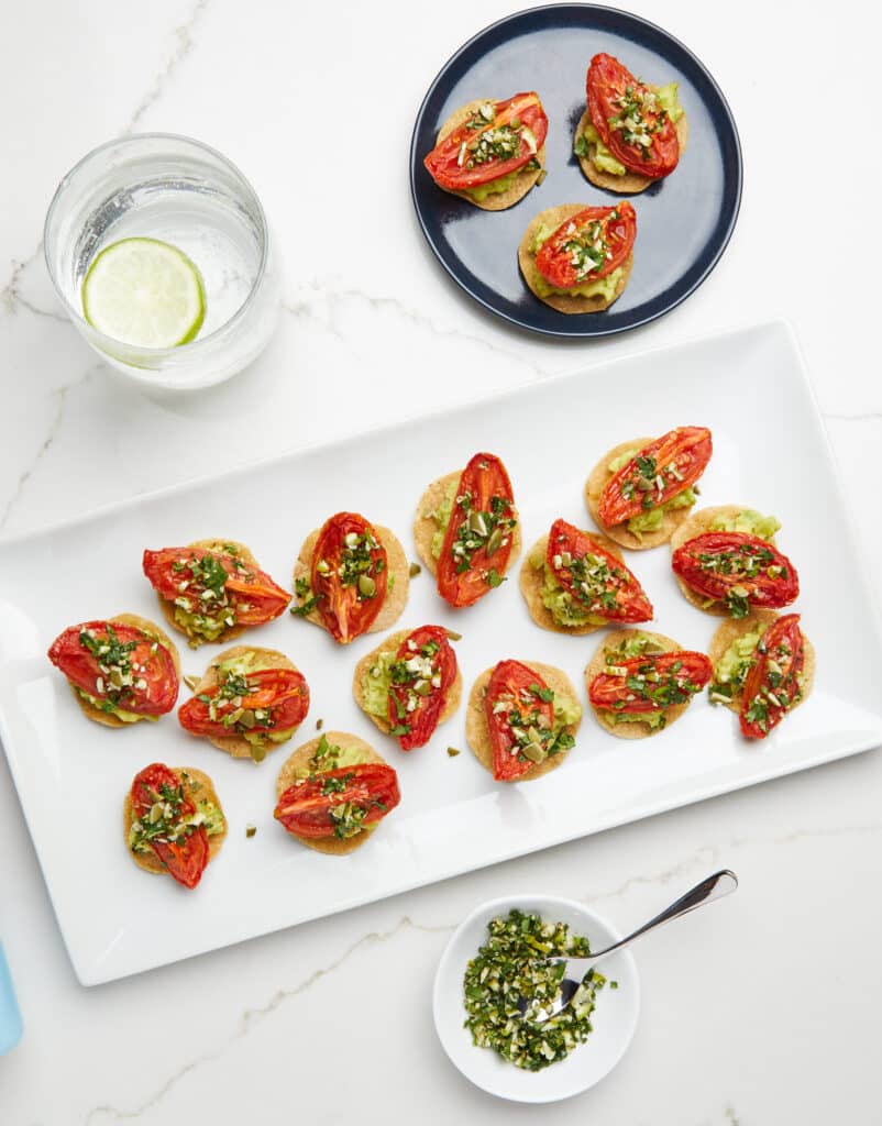 Colorful platter of roasted tomato and avocado appetizers garnished with herbs, served alongside a small bowl of chopped green salsa and a glass of water with lime. Ideal for a healthy snack or party appetizer.