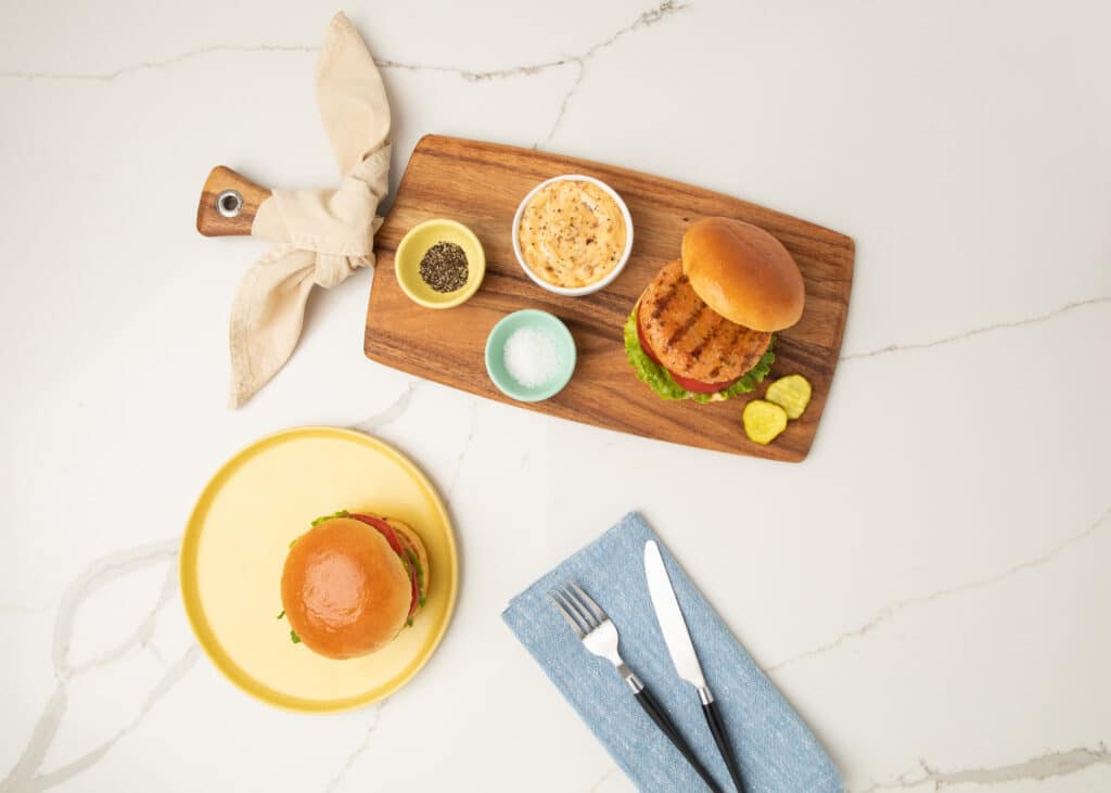 A delicious grilled shrimp and andouille sausage burger with lettuce and tomato on a wooden serving board, accompanied by pickles, seasoning, and a dipping sauce. A yellow plate with another chicken burger is neatly arranged beside a blue napkin and cutlery.