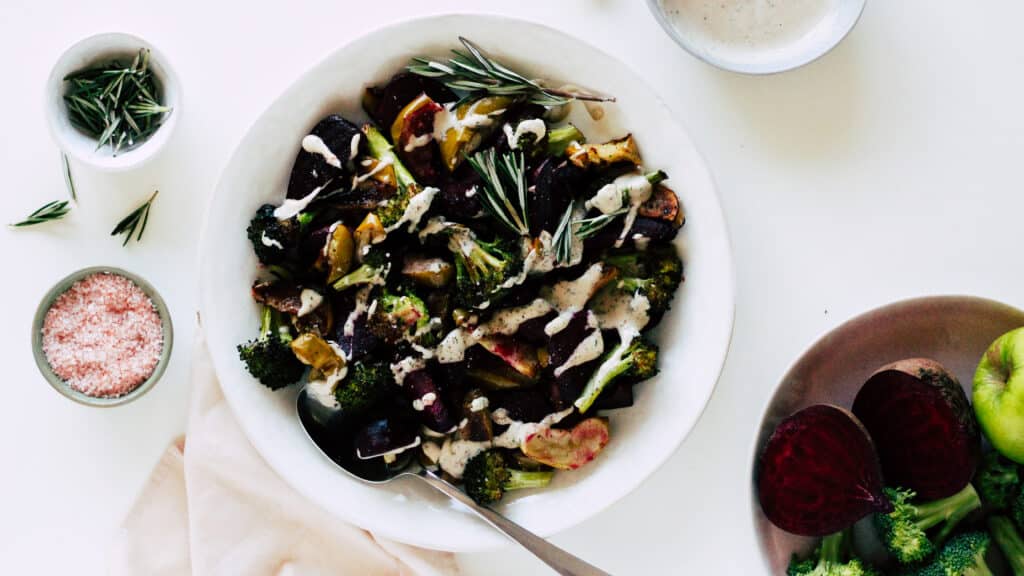 Colorful roasted vegetables including broccoli, beets, and apples drizzled with a creamy sauce, served in a white bowl, with fresh rosemary and pink salt on the side.