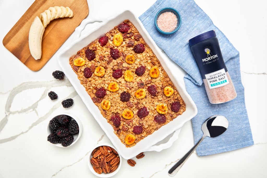 Baked oatmeal topped with sliced bananas, blackberries, and pecans, accompanied by a container of Morton Himalayan pink salt and a spoon, set on a marble surface with a wooden cutting board and additional blackberries.