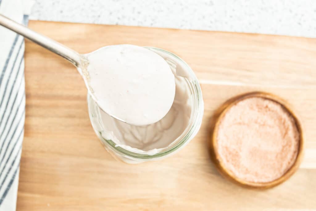 Creamy tahini sauce in a glass jar with a spoonful lifted above, accompanied by a small wooden bowl of pink salt, set on a wooden cutting board.