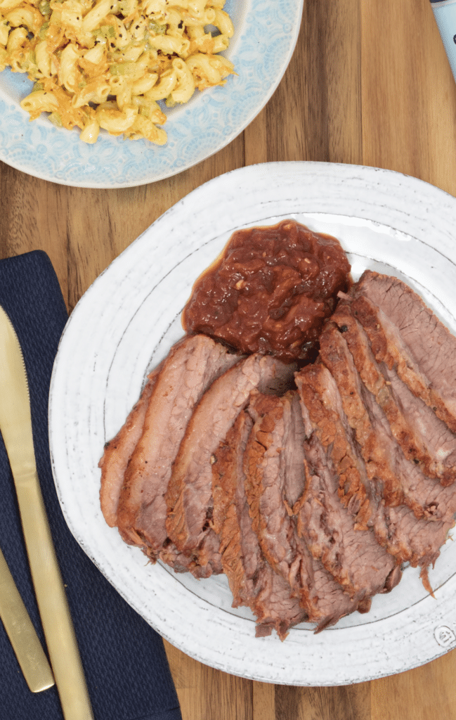 Sliced roast beef served with a side of tangy barbecue sauce, accompanied by a plate of creamy macaroni salad, on a wooden table setting with gold cutlery and a navy napkin.
