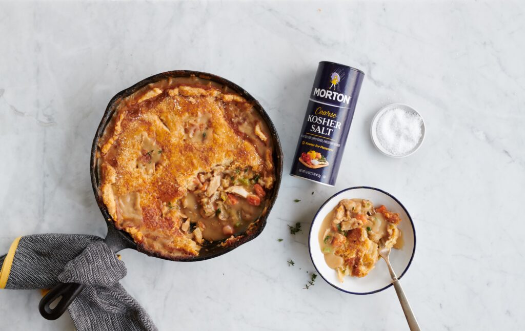 Skillet chicken pot pie with a golden crust, served in a cast iron skillet alongside a bowl of creamy filling and a can of Morton coarse kosher salt, set on a marble countertop.