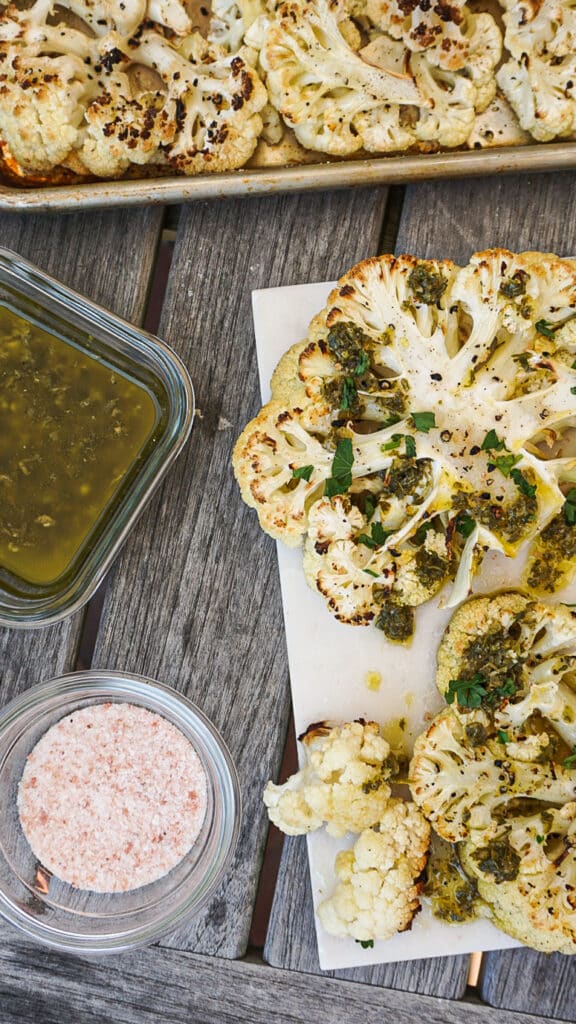 Roasted cauliflower steaks topped with green sauce and fresh herbs, served with a bowl of pink salt and a container of sauce, on a wooden table. Ideal for healthy vegetarian meals.