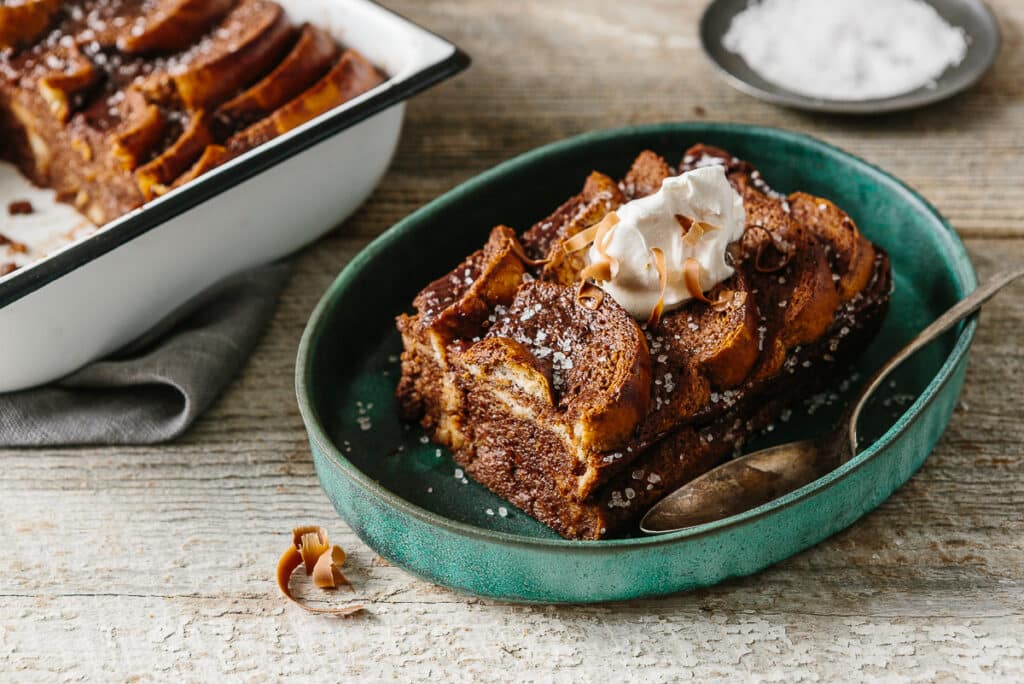 Delicious baked French toast served on a green plate, topped with whipped cream and chocolate shavings, with a sprinkle of sea salt, next to a baking dish on a rustic wooden surface.