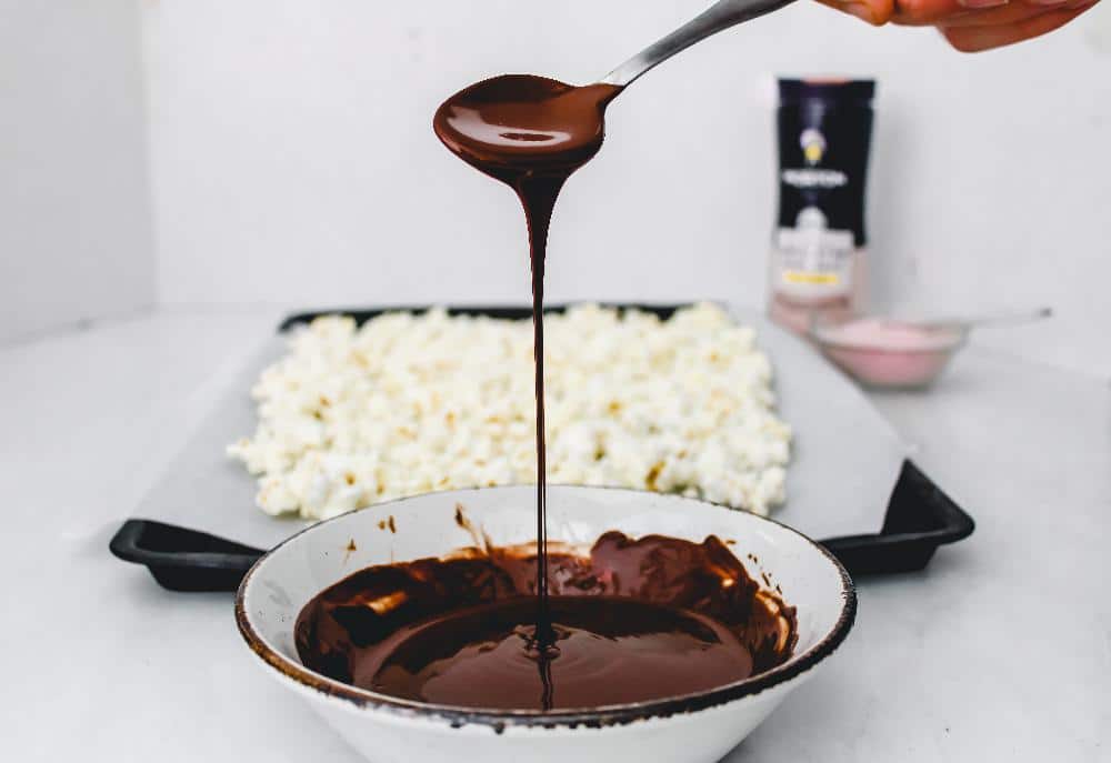 Bowl of melted chocolate with a spoon dripping chocolate, in front of a tray of popcorn, showcasing a delicious snack preparation.