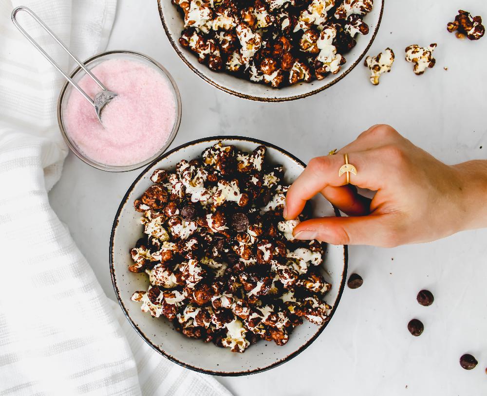 A hand reaching for a bowl of chocolate-covered popcorn, with a side bowl of pink salt, on a marble surface. The image showcases a delicious snack perfect for movie nights or parties.