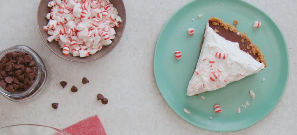 Slice of peppermint chocolate pie on a teal plate, surrounded by crushed peppermint candies in a bowl and chocolate chips scattered on a light surface, perfect for holiday dessert inspiration.