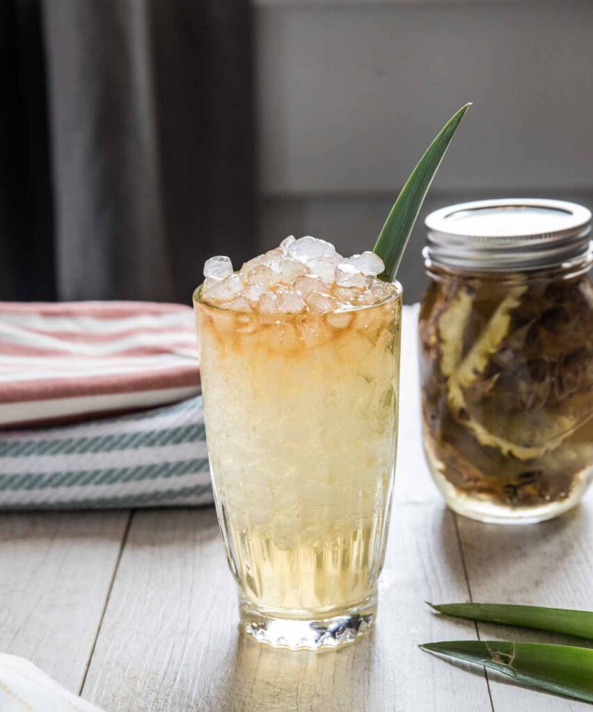 Refreshing cocktail served in a tall glass filled with crushed ice, garnished with a green leaf, alongside a jar of preserved ingredients and colorful kitchen linens. Perfect for summer gatherings or tropical-themed parties.