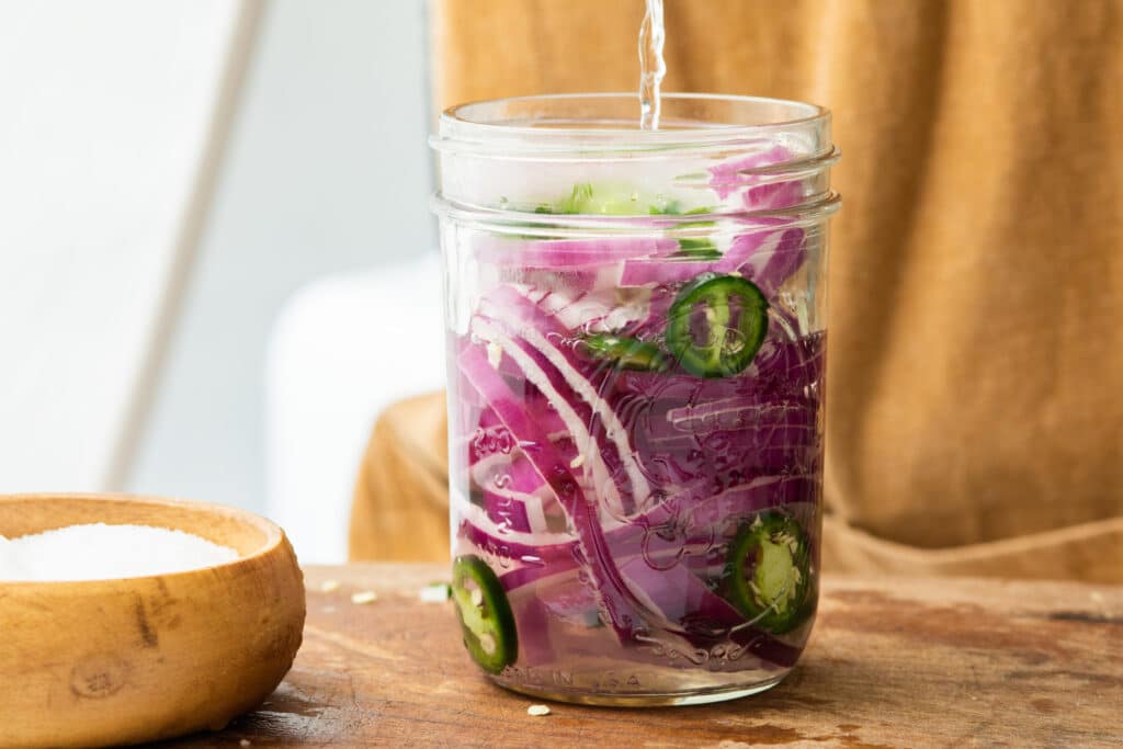 Jar of pickled red onions and jalapeños being prepared with water, placed on a wooden surface next to a bowl of salt.