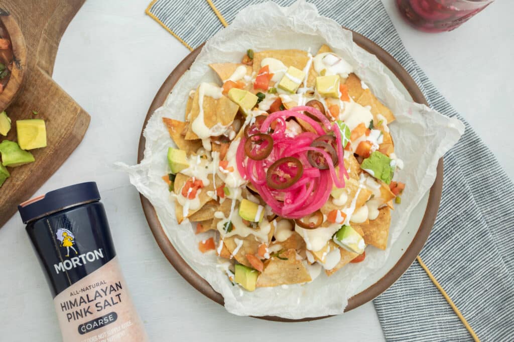 Delicious plate of nachos topped with creamy sauce, diced tomatoes, avocado, pickled onions, and jalapeños, accompanied by Morton Himalayan pink salt on the side. Perfect for sharing at parties or enjoying as a snack.