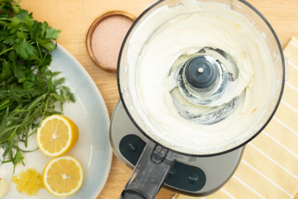 Close-up of a food processor filled with creamy white sauce, surrounded by fresh herbs, sliced lemons, and a small bowl of salt on a wooden countertop. Ideal for food preparation and cooking inspiration.