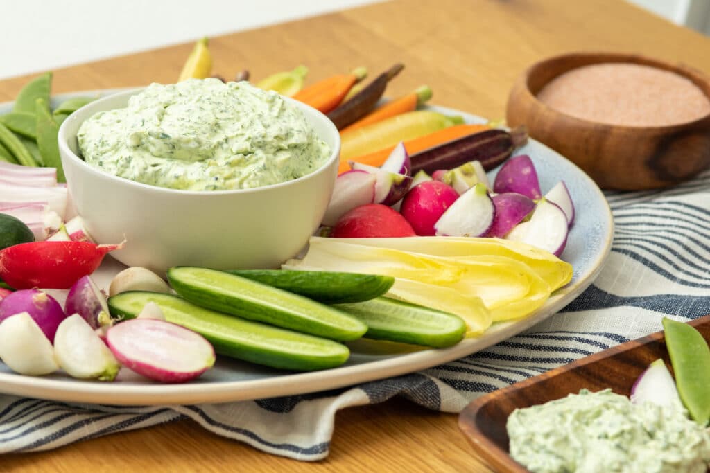 Fresh vegetable platter featuring colorful radishes, cucumbers, and carrots, accompanied by a creamy herb dip in a white bowl. The arrangement is set on a wooden table with a striped cloth, perfect for healthy snacking or entertaining.