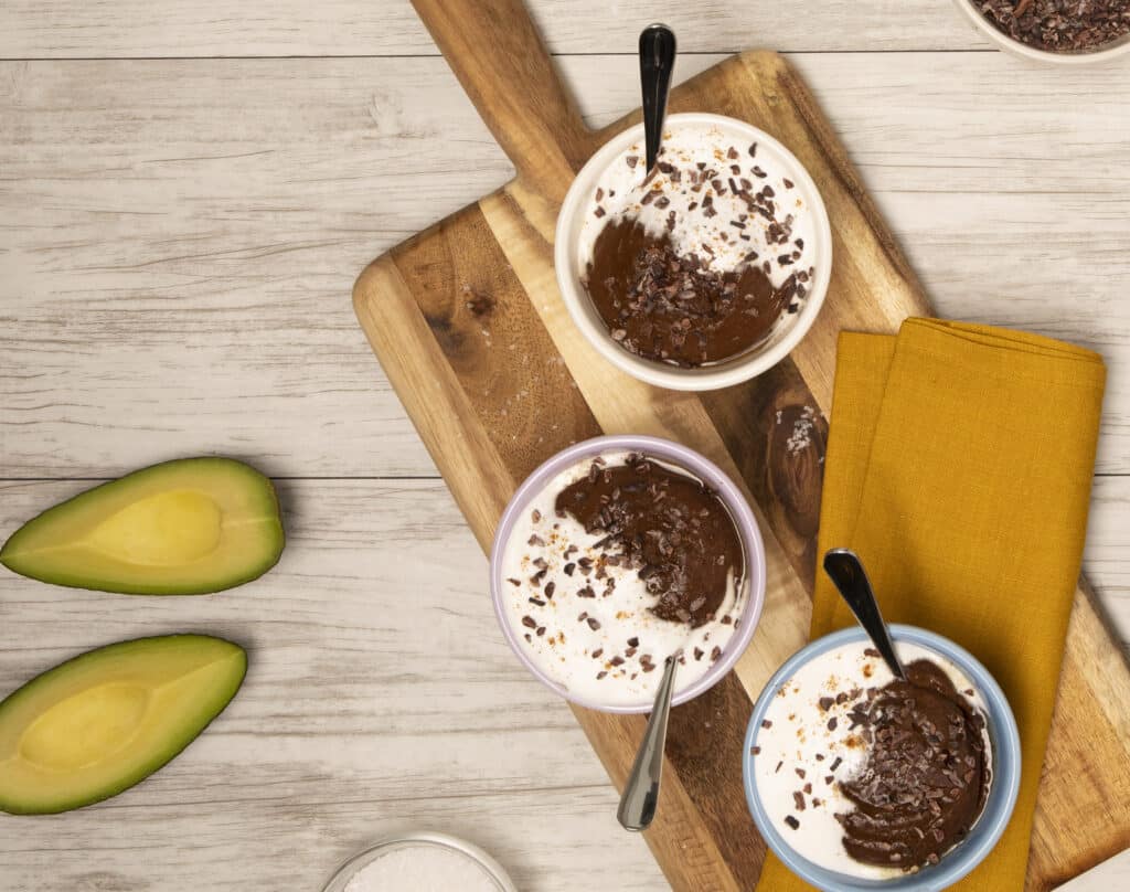 Delicious avocado chocolate mousse served in colorful bowls, topped with whipped cream and cacao nibs, on a wooden cutting board with fresh avocado slices and a yellow napkin.