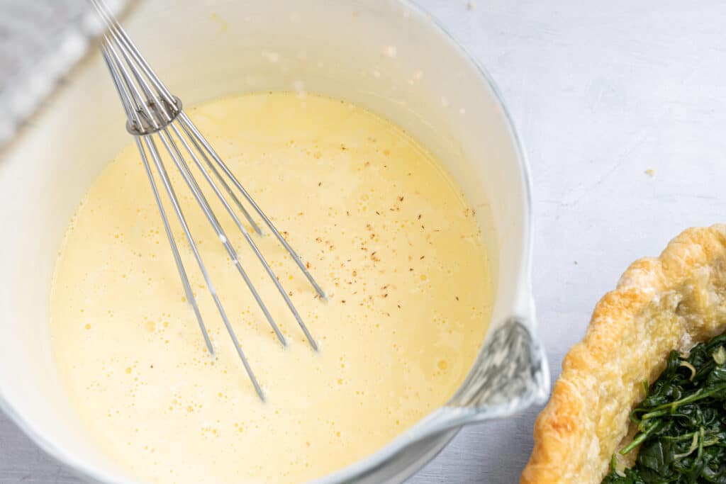 Bowl of whisked egg mixture with seasoning, preparing for a savory quiche, alongside a pie crust filled with cooked spinach.